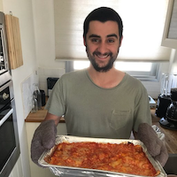 The founder, grinning, holding a freshly baked tray of lasagna in oven mitts